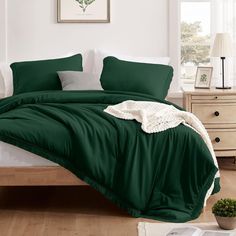 a bed with green sheets and pillows in a room next to a white dresser, potted plant and window