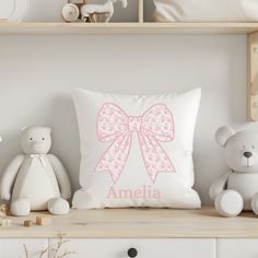 a white pillow with a pink bow on it sitting next to two stuffed animals and a teddy bear