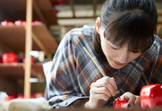 a woman in plaid shirt painting an apple with red paint on the table next to shelves