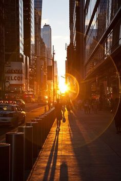 the sun is setting on a city street with tall buildings and people walking down the sidewalk