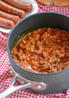 chili in a pot next to hot dogs on a checkered tablecloth with red and white napkins