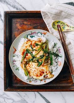 a white plate topped with chicken and rice next to chopsticks on a wooden tray