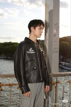 a young man wearing a black leather jacket and striped pants standing next to a body of water