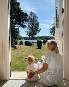 a woman sitting on the floor with her child looking out an open door at a lake