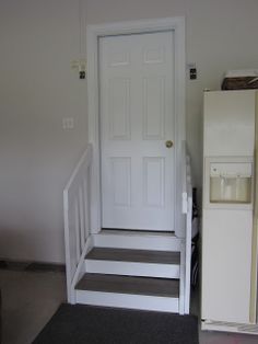 a white refrigerator sitting next to a door in a room with stairs leading up to it