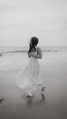 black and white photograph of woman in flowing dress walking on the beach with waves coming in