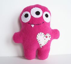 a pink stuffed animal with big eyes and a heart on it's chest sitting against a white wall