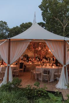 an outdoor tent with tables and chairs set up for a wedding reception at dusk, surrounded by greenery