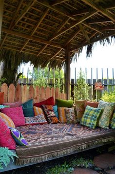 a couch covered in lots of pillows sitting under a wooden roof next to a fence