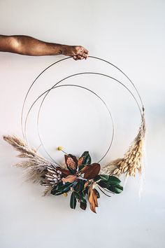 a person is holding a circular wreath with dried flowers and leaves on the side, while another hand holds it up