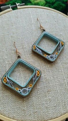 two square shaped earrings sitting on top of a piece of fabric next to each other