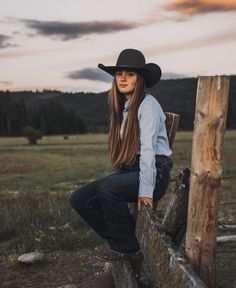 a beautiful young woman wearing a cowboy hat leaning on a wooden fence in a field