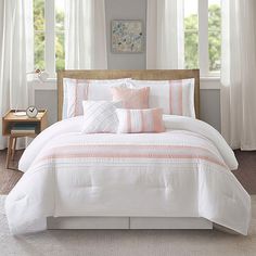 a bed with white and pink pillows in a bedroom next to two large windows,