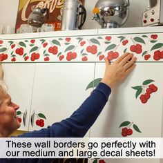 an older woman is painting the wall with cherry decals on her kitchen cupboards