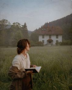 a woman standing in the middle of a field holding a book and looking at a house