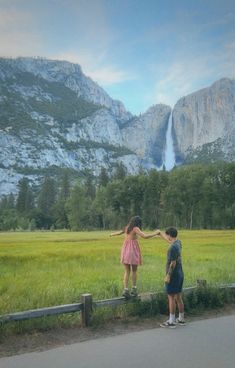 two people standing on the side of a road in front of a mountain with a waterfall