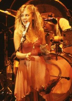 a woman in a pink dress holding a microphone and standing next to a drum set