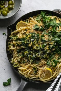 a skillet filled with pasta and pesto on top of a white table next to olives