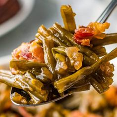 a spoon full of cooked green beans with bacon on top and another dish in the background