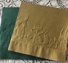 two napkins with animals on them sitting on a lace doily covered tablecloth