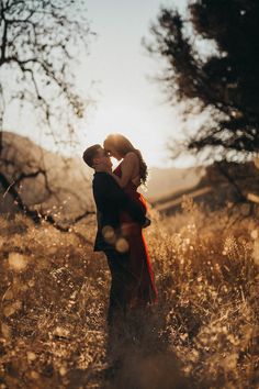 a man and woman standing in tall grass with the sun shining through trees behind them