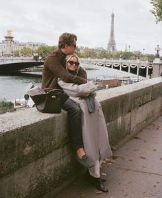 a man and woman sitting next to each other on a wall near the water with eiffel tower in the background