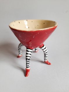 a red bowl with black and white striped legs on it's legs, sitting in front of a gray background