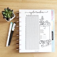 an open planner sitting on top of a wooden table next to a pen and potted plant