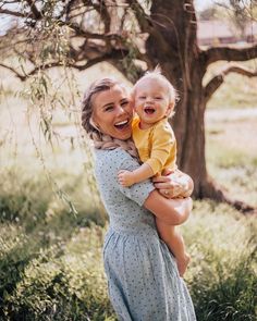 a woman holding a baby in her arms and smiling at the camera while standing next to a tree