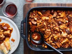 a casserole dish with meat and vegetables on a wooden cutting board next to wine glasses