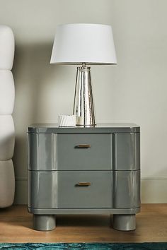 a nightstand with a lamp on top of it next to a white chair and ottoman