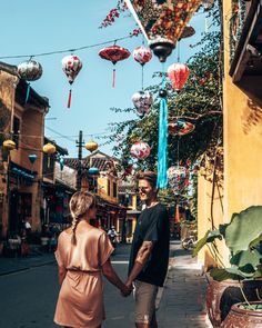 a man and woman holding hands walking down the street with lanterns hanging from the buildings