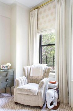 a white chair sitting in front of a window next to a dresser with drawers on it