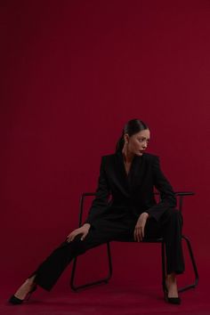 a woman is sitting on a chair in front of a red background wearing a black suit