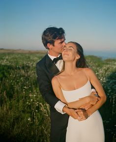 a bride and groom kissing in a field with the ocean in the background