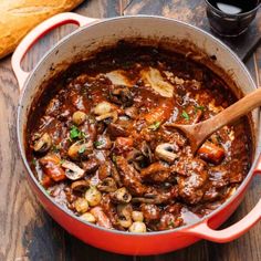 a pot filled with stew and mushrooms on top of a wooden table next to bread