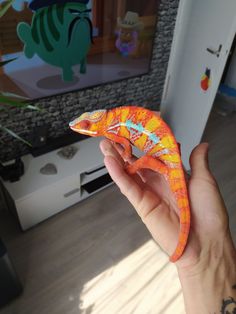 a hand holding an orange gecko toy in front of a television set with cartoon characters on it