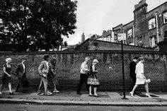 black and white photograph of people walking on sidewalk next to brick wall in urban area