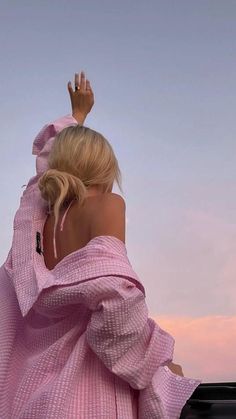 a woman in a pink dress standing on the roof of a car with her hand up