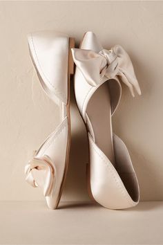 a pair of white wedding shoes with a bow on the toe and heel, against a beige wall