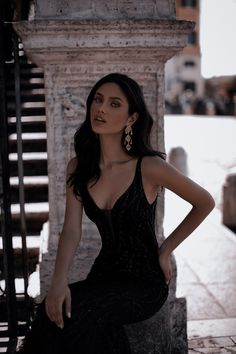 a woman in a black dress leaning against a stone wall with her hands on her hips