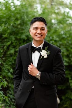 a man in a tuxedo smiles at the camera while standing next to some bushes