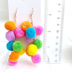 multicolored pom pom - pom earrings hanging from gold chain, next to measuring ruler