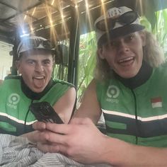 two men in green vests sitting next to each other on a bed and one is holding a cell phone