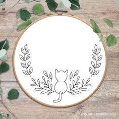 an embroidery hoop with leaves and a cat in the center on a wooden surface, surrounded by greenery