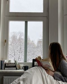 a woman laying in bed reading a book next to a window with snowy trees outside