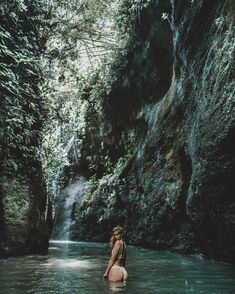 a woman standing in the middle of a river next to a lush green forest filled with trees