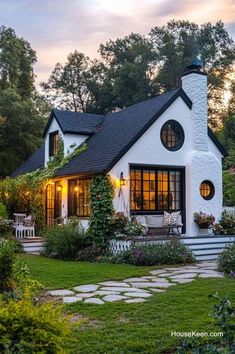 a white house with black roof and windows in the front yard at dusk, surrounded by greenery