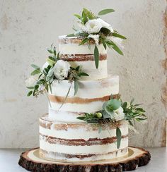 a three tiered cake with white flowers and greenery on the top is sitting on a wood slice