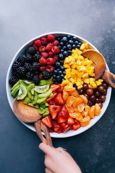 a bowl filled with fruits and vegetables on top of a table next to a person's hand